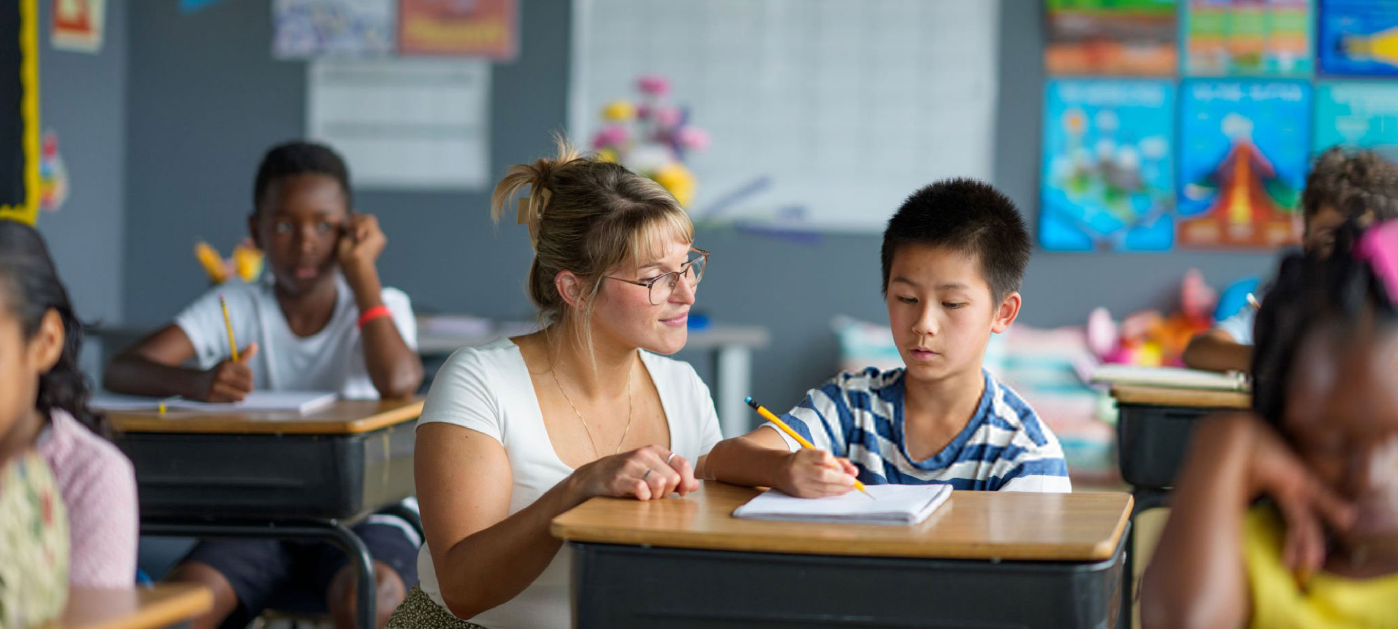 A female teacher crouches beside a desk explaining the lesson to her pupil.